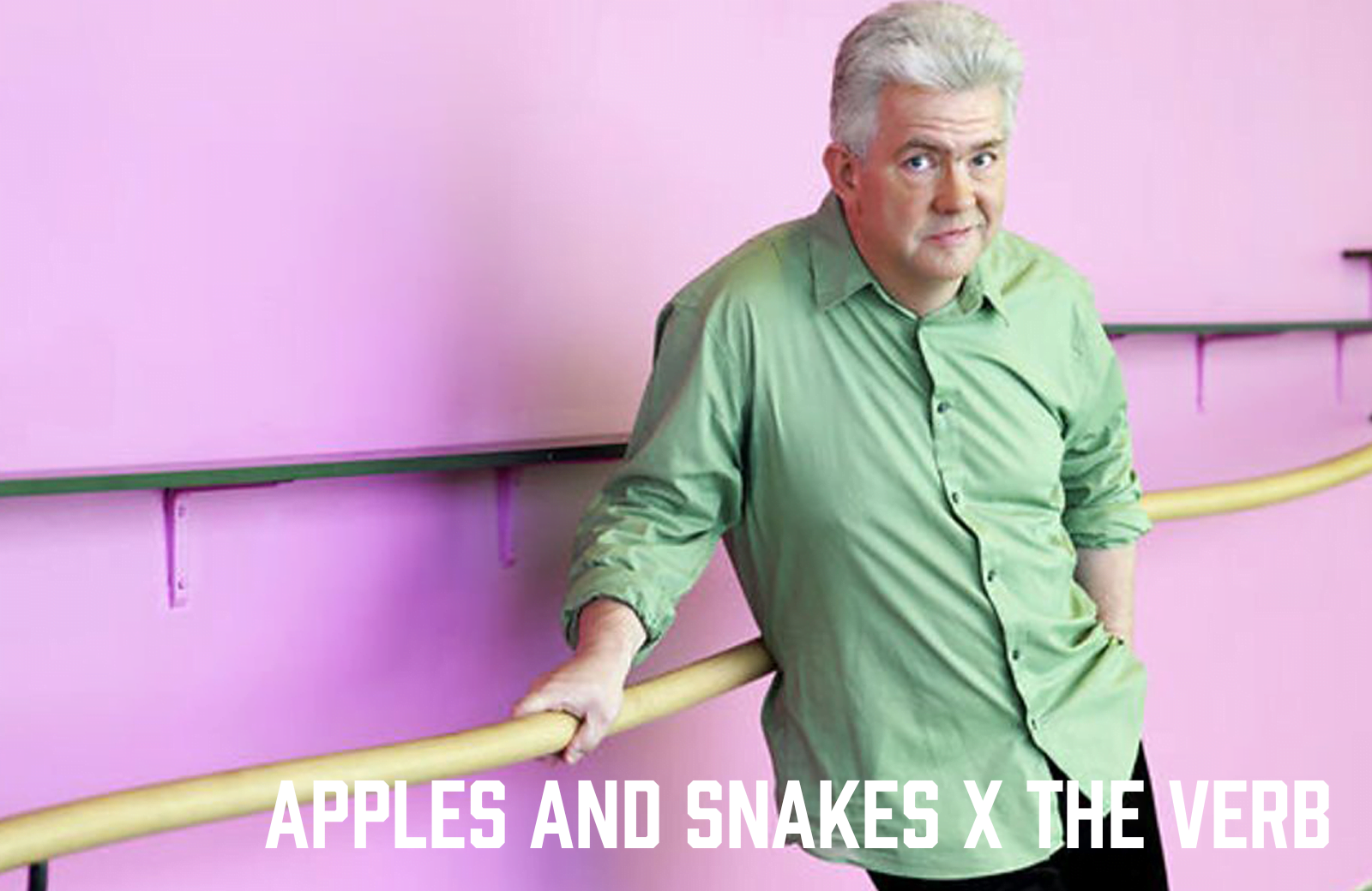 A grey haired man in a green shirt, leans against a cream banister against a pink wall